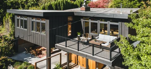 A modern two-story home with dark charcoal siding, a large raised balcony, and a ground-level patio nestled in trees.