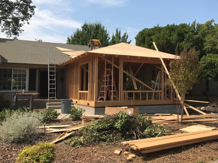 A construction site shows a wooden home addition frame attached to the side of an existing house with a roof partly laid.