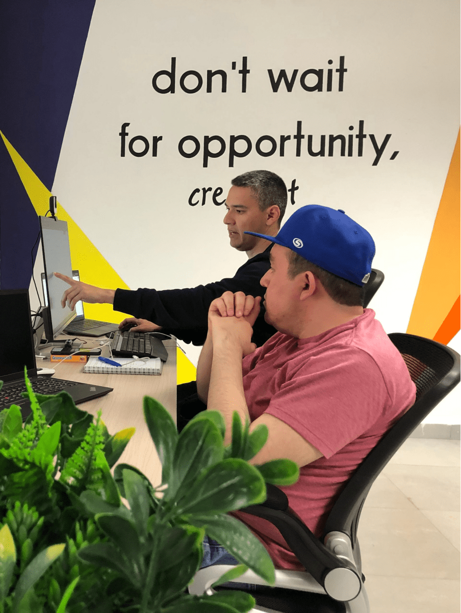 Two men are sitting at a desk in front of a wall that says 