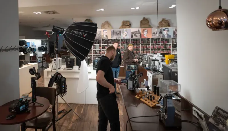 Set einer Videoproduktion: Menschen in einem Geschäft für Kaffeemaschinen, in dem die Lichtanlage für ein Fotoshooting aufgebaut ist. Ein Videograf steht an einem Tresen.