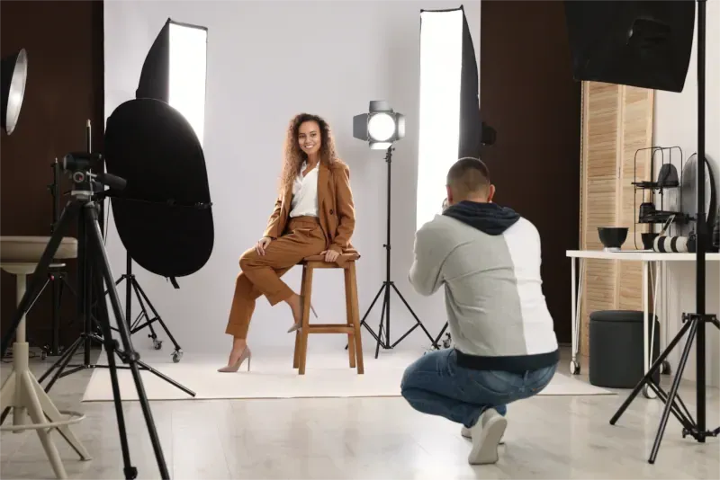 Ein Fotograf fotografiert eine Frau in einem braunen Anzug, die in einem Studio mit Scheinwerfern und Hintergründen auf einem Hocker sitzt.