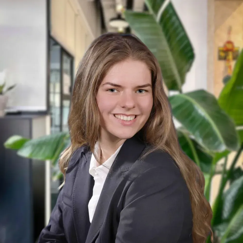 Eine lächelnde junge Frau in einem dunklen Blazer und einem weißen Hemd sitzt in einem Innenraum vor dem Hintergrund grüner Pflanzen.