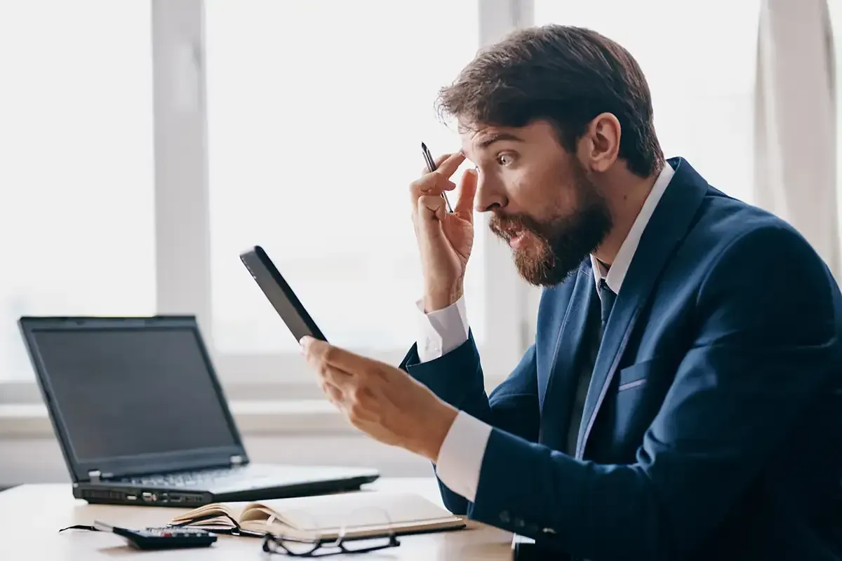 Ein Mann im Anzug sitzt an einem Schreibtisch, schaut überrascht und hält ein Tablet in der Hand. Vor ihm steht ein Laptop, die Szene spielt in einem Büro.