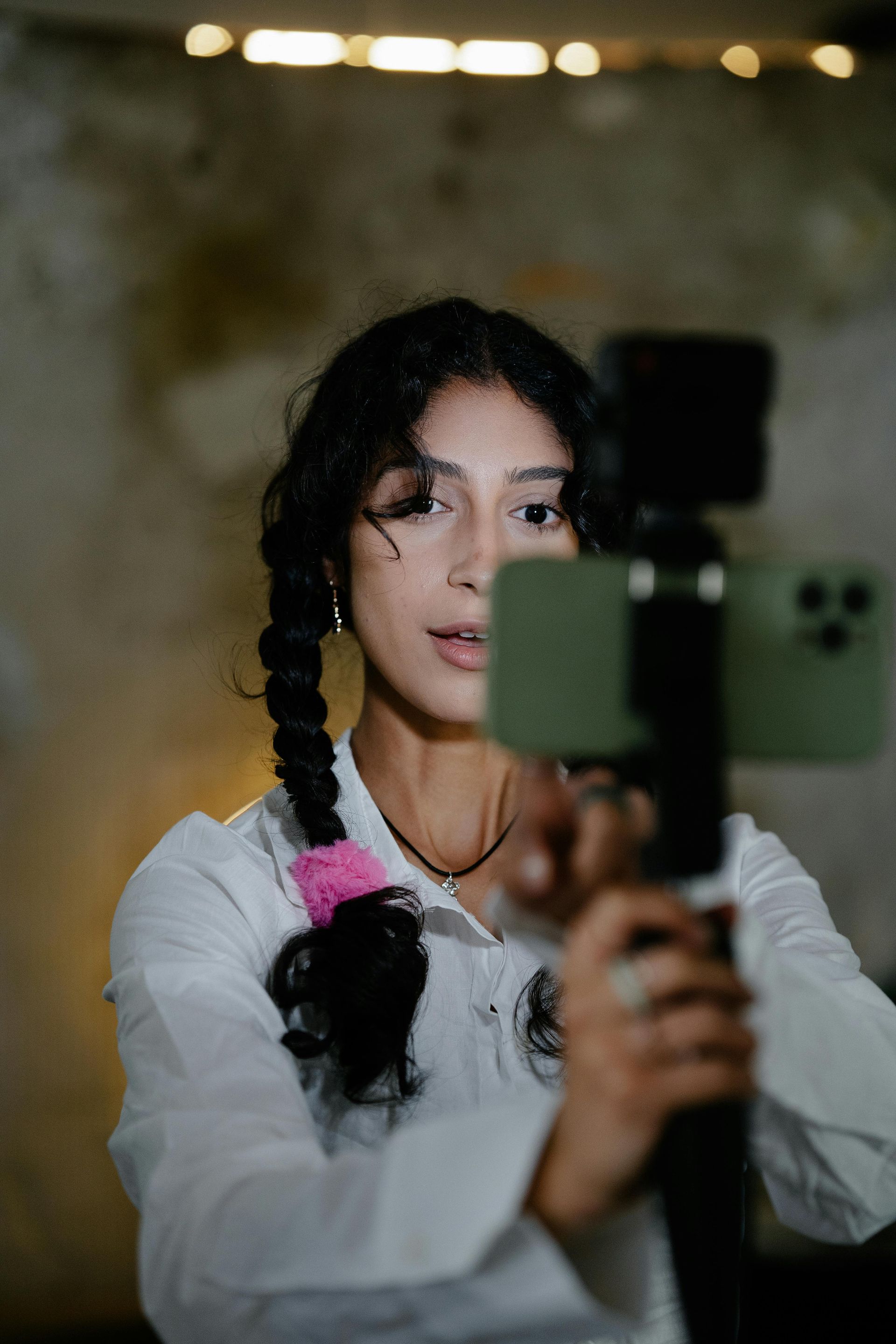 Eine Frau, die gerade ein Reel mit einem Smartphone und einem Gimbal produziert, trägt ein weißes Hemd und ein rosa Haargummi.