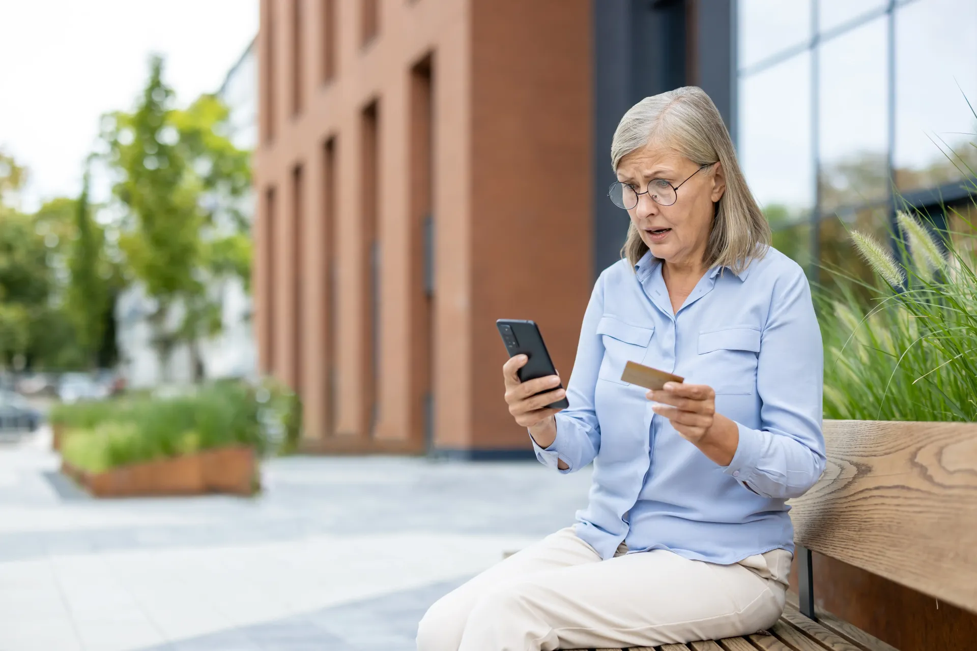 Eine ältere Frau sitzt auf einer Bank und hält eine Kreditkarte und ein Mobiltelefon in der Hand.