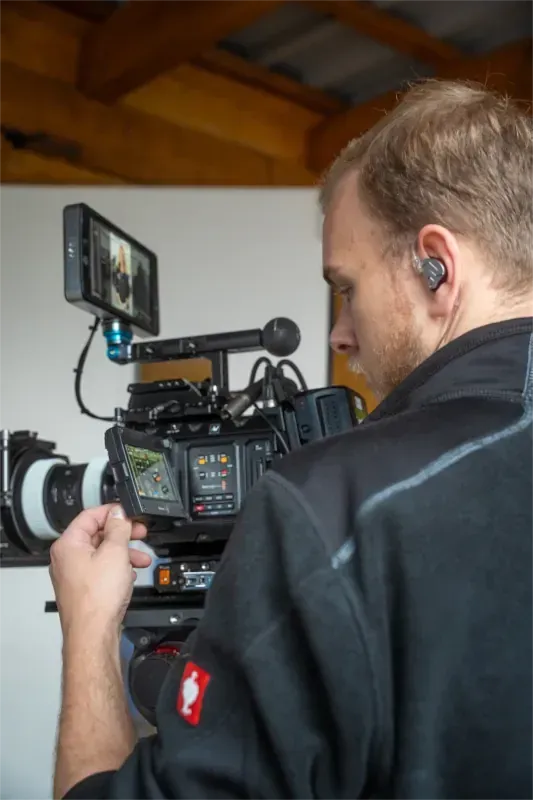 Ein Filmemacher bedient eine Videokamera und schaut dabei auf den Bildschirm – alles in Innenräumen.