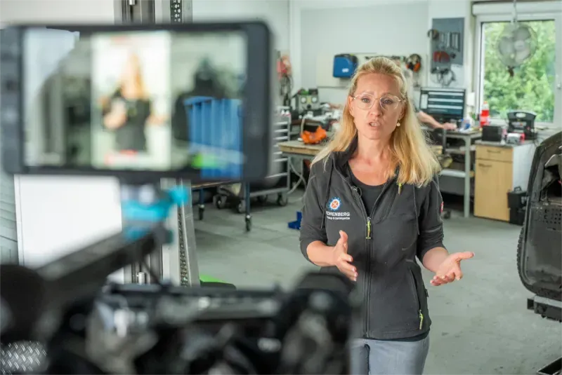 Frau in der Garage, spricht in die Kamera. Filmausrüstung im Vordergrund, Werkzeuge und Arbeitsbereich im Hintergrund.