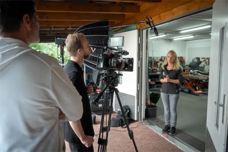 Eine Frau wird in einem Garagentor gefilmt; das Filmteam ist mit Kameras und Mikrofonen ausgestattet.