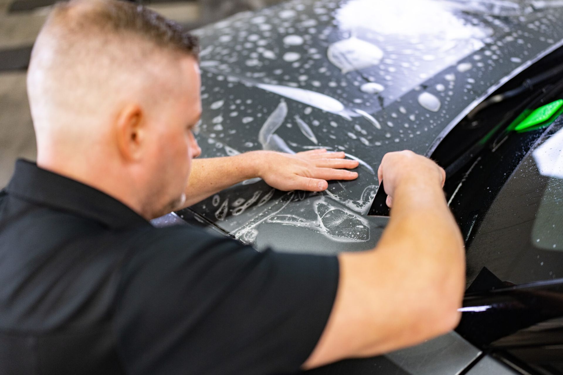 Person applying a clear protective film to a black car hood in a workshop
