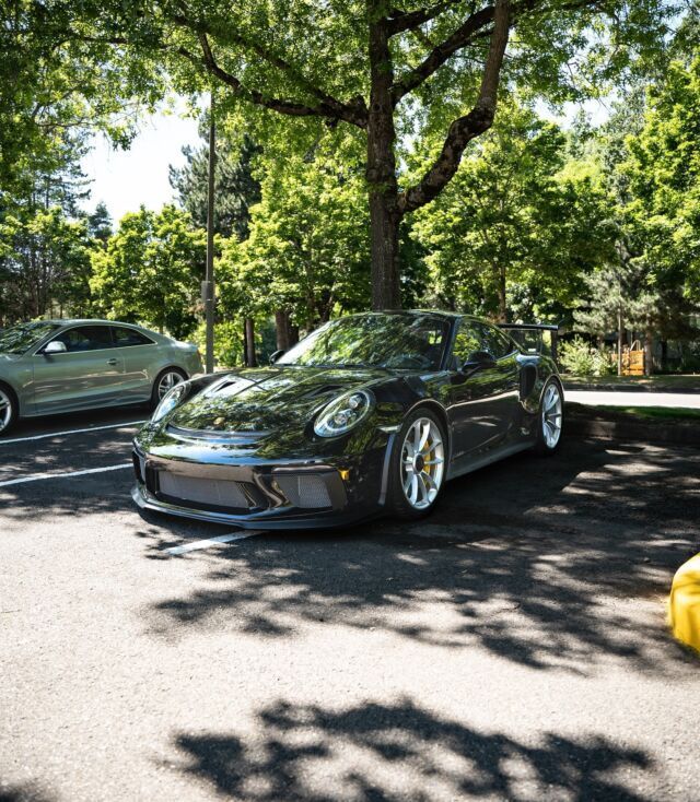 Black sports car parked under trees in a sunny lot, with another car nearby.