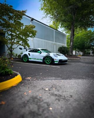 White sports car with green rims parked beside a gray building under trees.