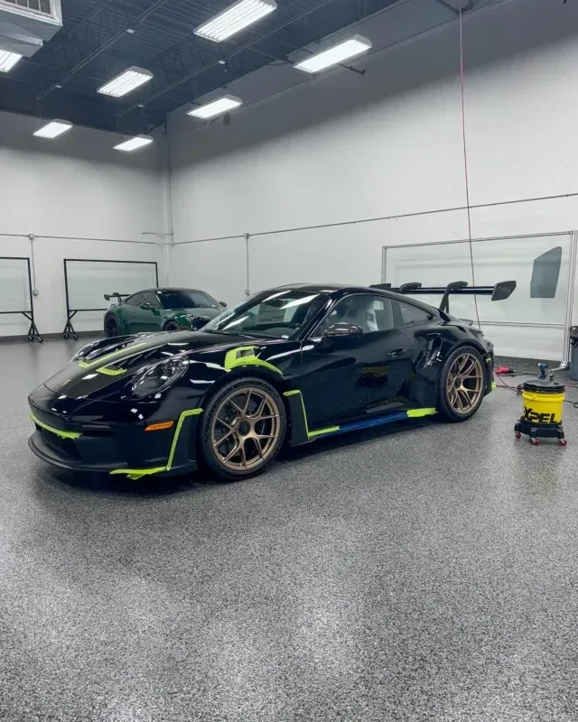 Black sports car with neon yellow accents in a garage, next to cleaning equipment and another car