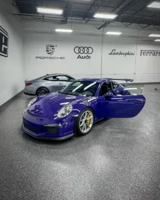 Purple Porsche in a showroom with luxury car brand logos on the wall and a silver car behind it