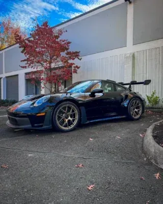 Blue sports car with rear wing parked outside a building with autumn trees nearby