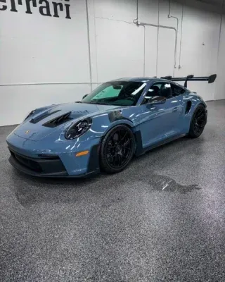 Blue Porsche sports car in a showroom, angled front-left view, with black wheels and a large rear wing