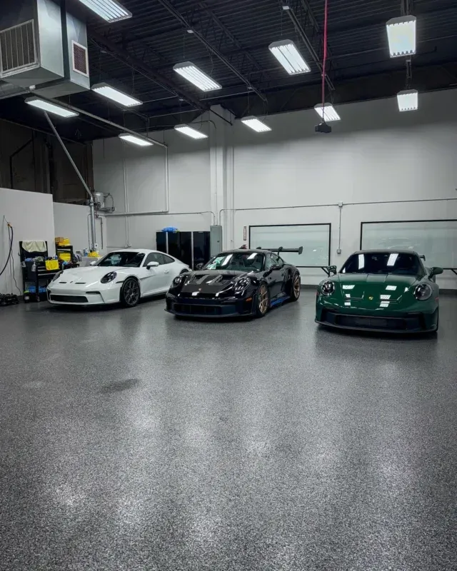 Three sports cars parked in a bright indoor garage: white, black, and green.