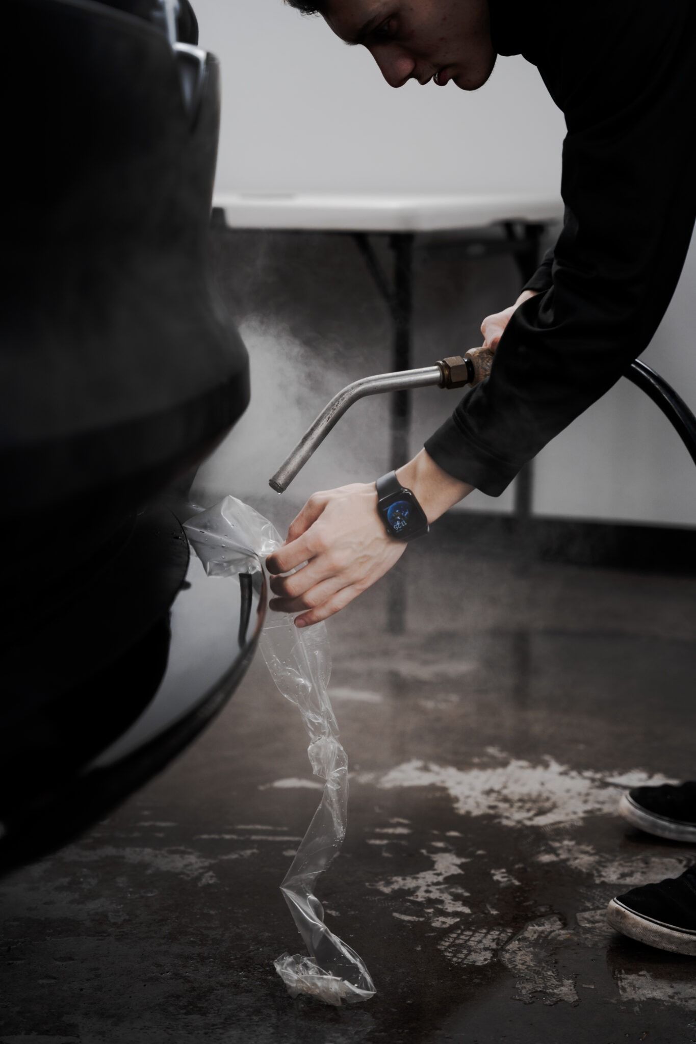 Person rinsing a black car tire with a hose, water splashing onto the wet floor.