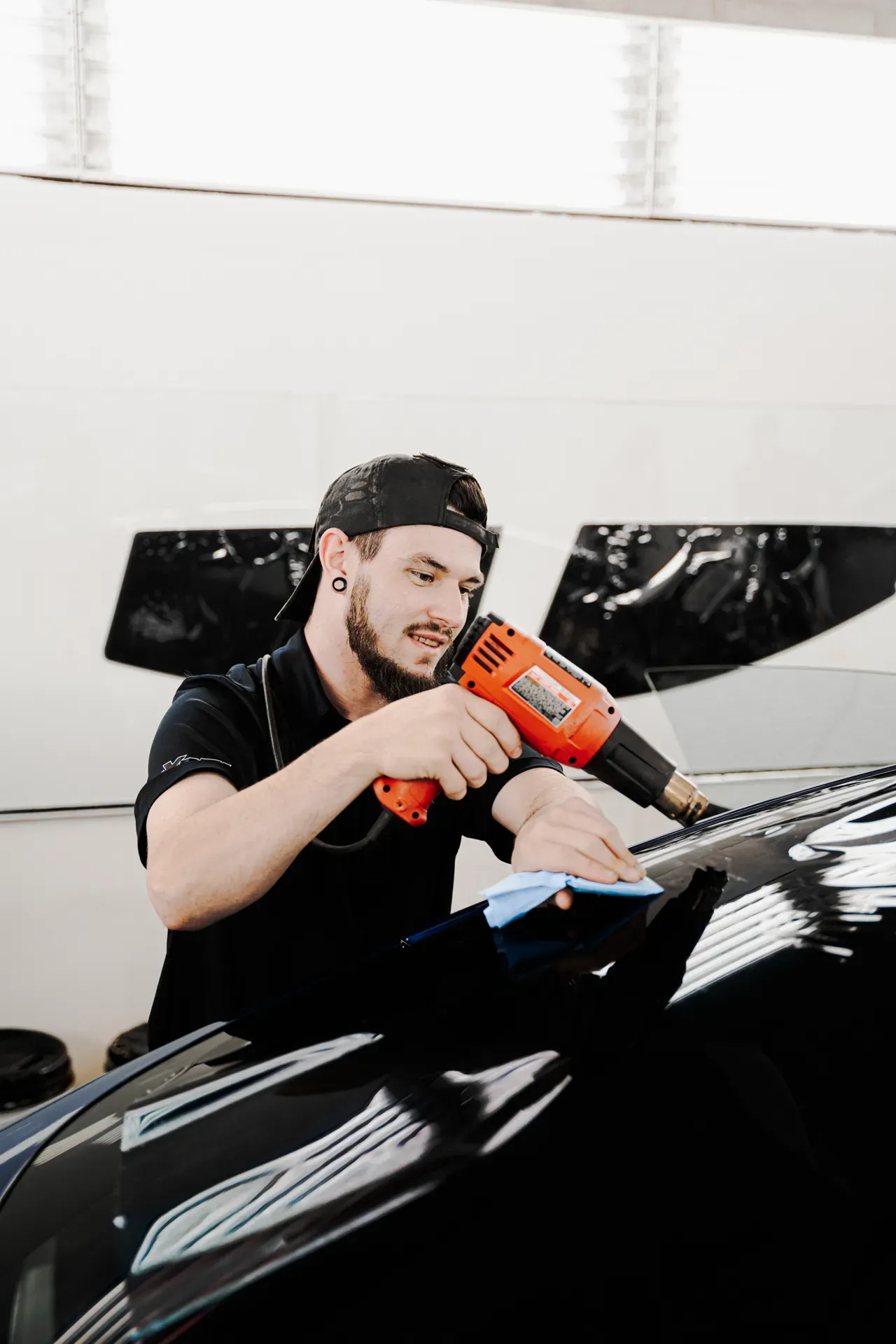 Mechanic using an orange heat gun to work on a glossy black car in a bright workshop