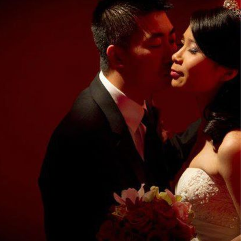 Bride and groom sharing a close kiss in a red-lit studio, holding a bouquet