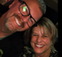 Two smiling people posing closely together in a dim setting, one wearing glasses.