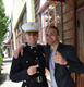 Two smiling men pose arm-in-arm outdoors, one in a dark uniform and white hat, the other in a dark blazer.