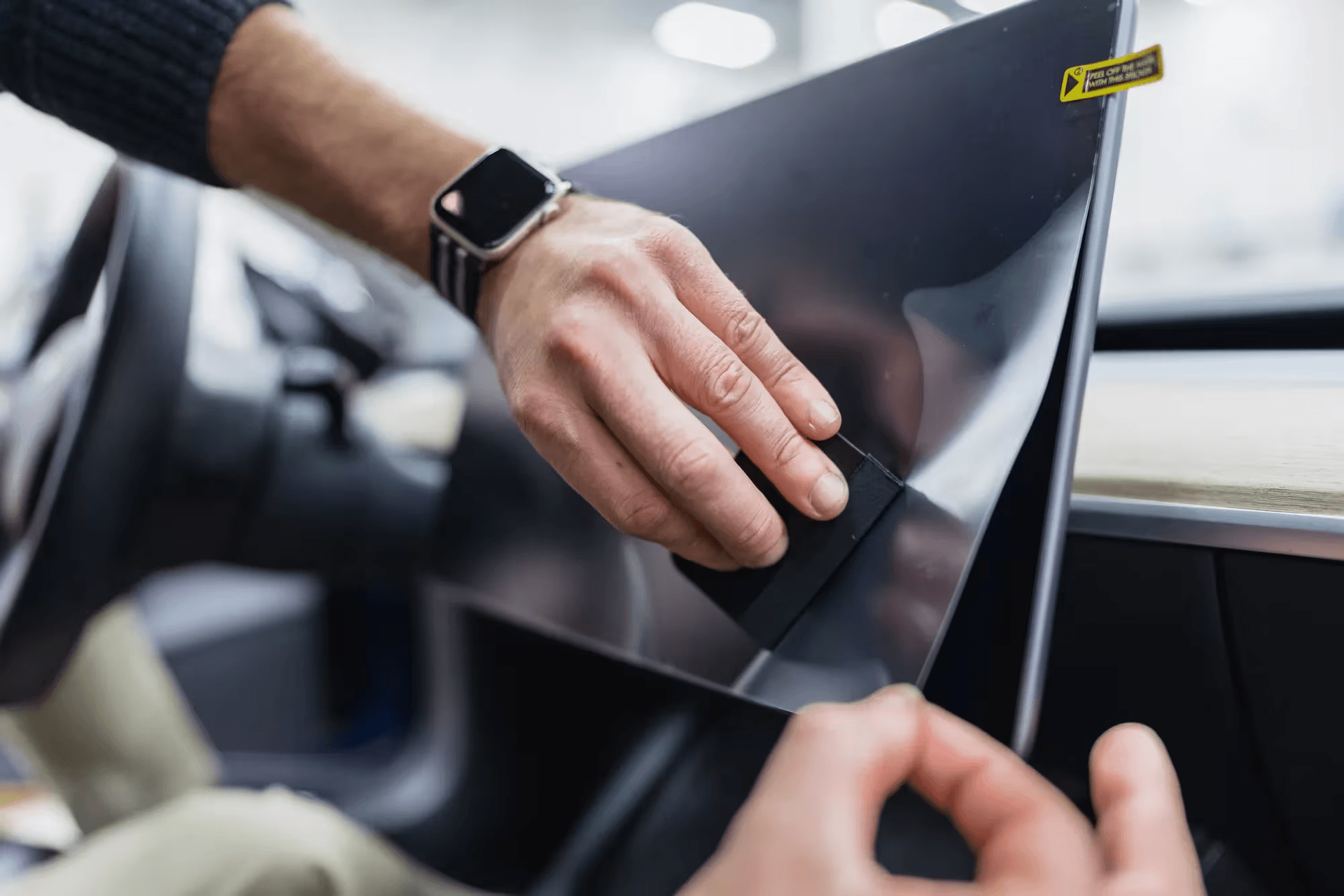 Hands peeling a clear protective film from a glossy black laptop or device screen in a car.