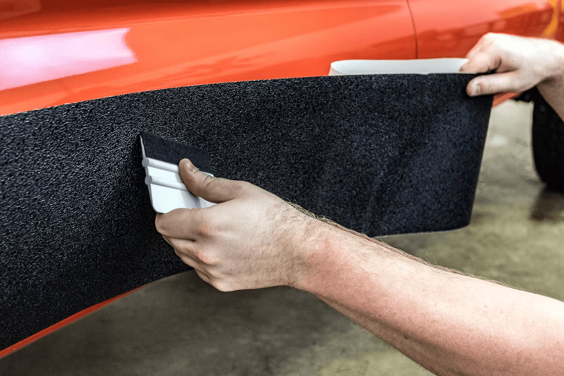 Hands installing a black side skirt on a red car in a garage.