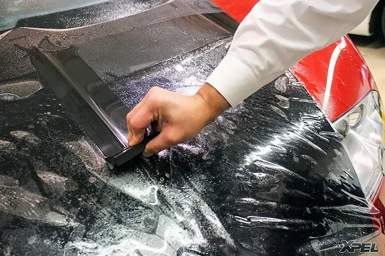 Person uses a squeegee on a black film covering a red car hood