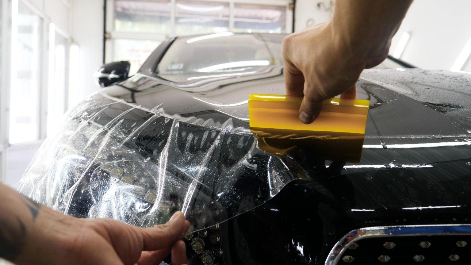 Hands squeegeeing a black car window with plastic wrap during detailing