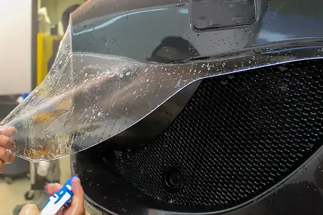 Peeling clear protective film from a black car hood and grille with a hand tool