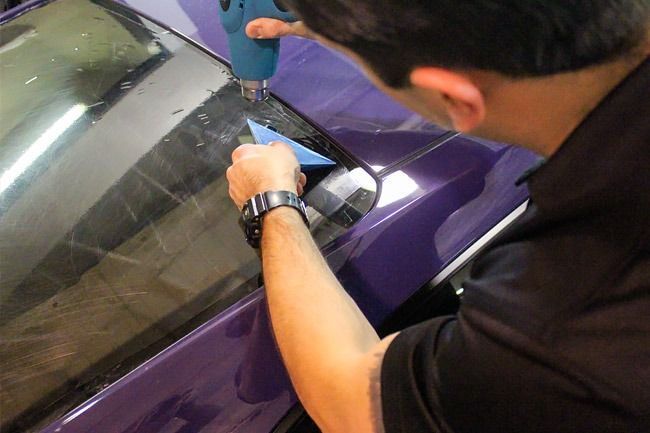 Person working on a purple car window with a blue tool in a shop