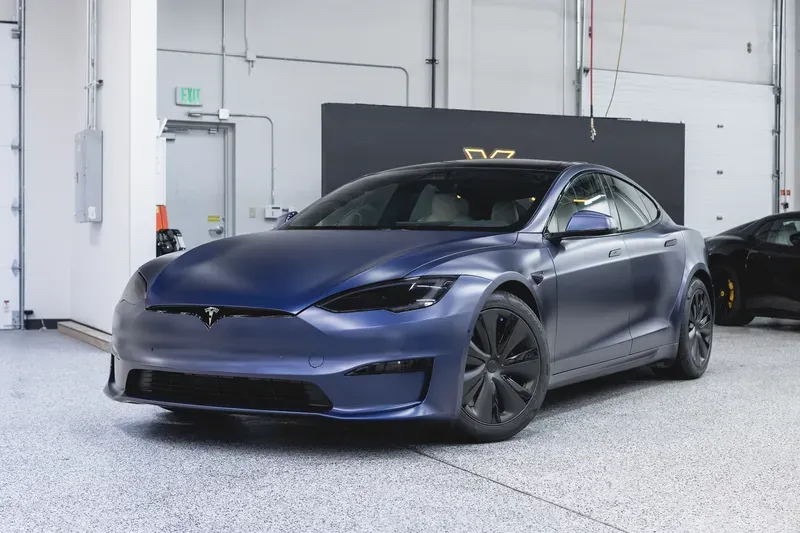 Blue Tesla sedan parked indoors in a bright garage with a white wall and gray floor