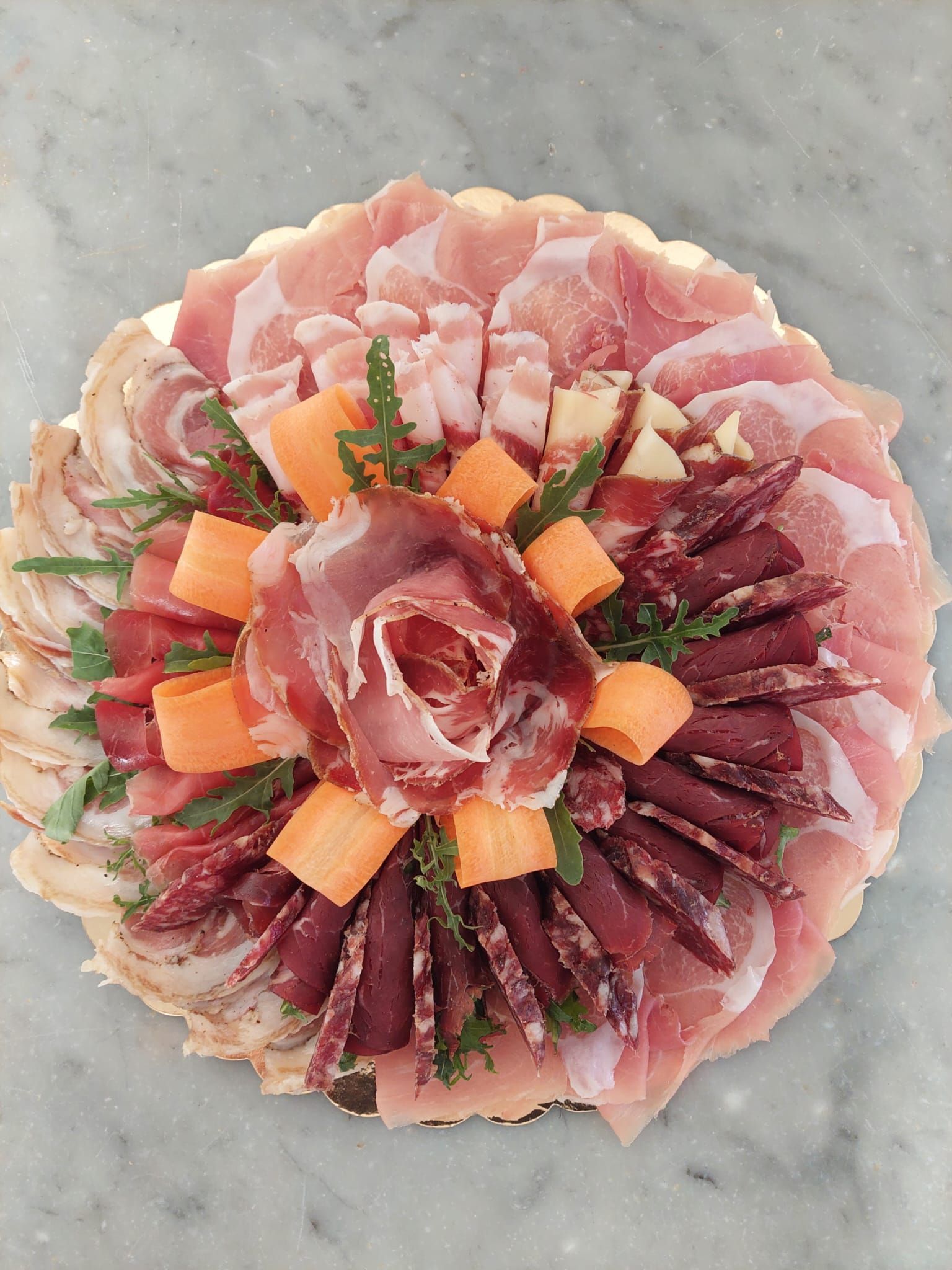 Pesce crudo assortito a fette, disposto a forma di fiore su un piatto grigio, guarnito con cubetti d'arancia ed erbe aromatiche.