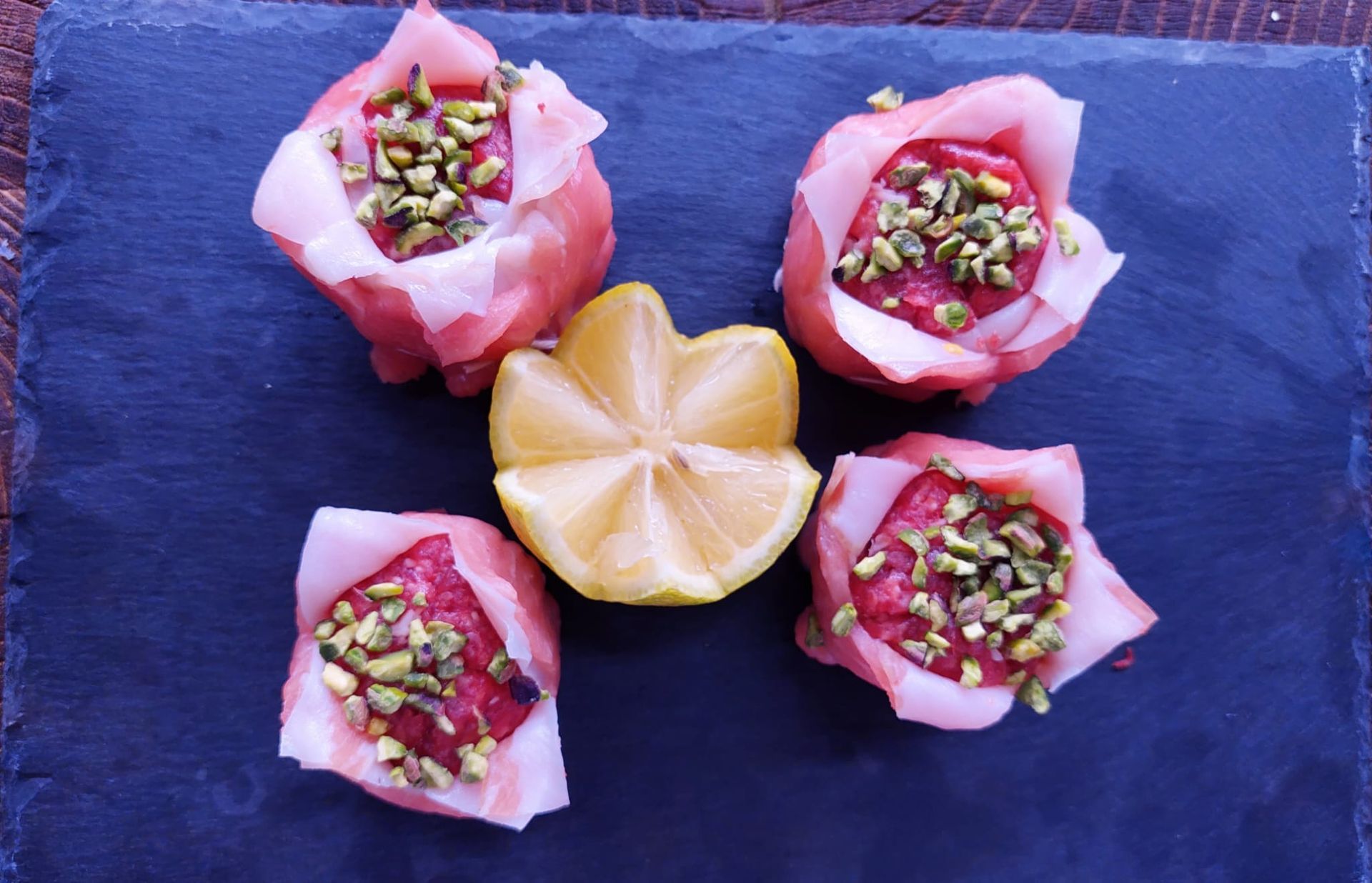 Pasticcini rosa a forma di rosa su una lastra di ardesia scura con una fetta di limone al centro, guarniti con frutta secca tritata.