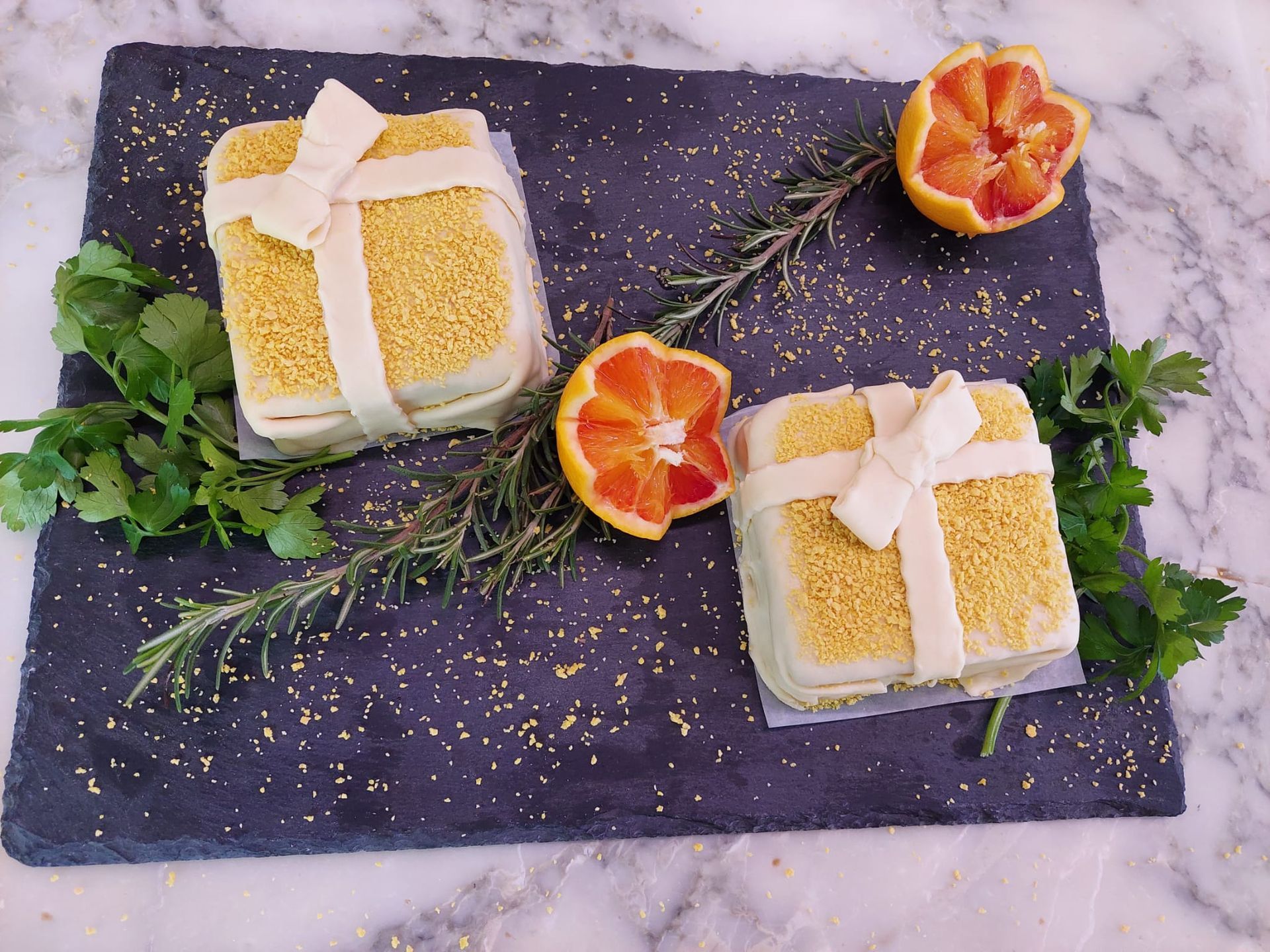 Due torte ricoperte di glassa bianca con fette di limone su un vassoio di ardesia scura, guarnite con erbe aromatiche e mezze arance.