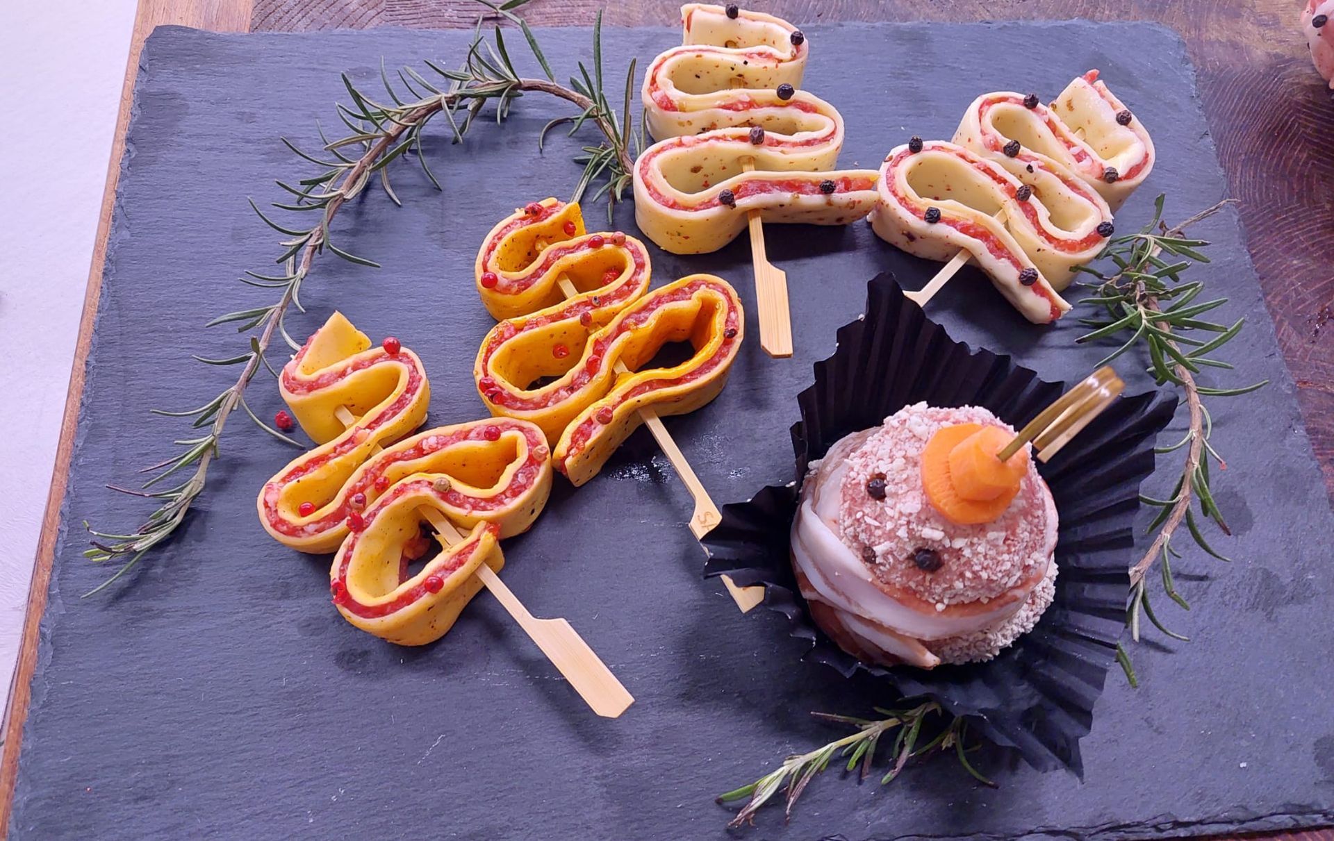 Cake pops colorati arrotolati su stecco con erbe aromatiche su un vassoio di ardesia accanto a un cupcake glassato.