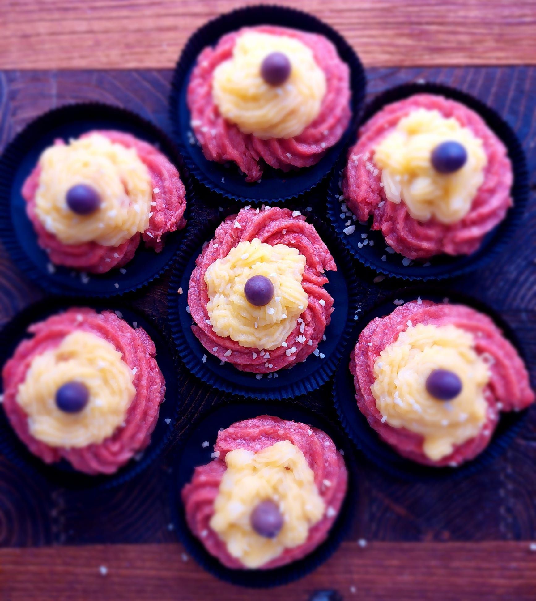 Assortimento di pasticcini simili a cupcake con glassa rosa e frutti di bosco, serviti su un vassoio scuro.