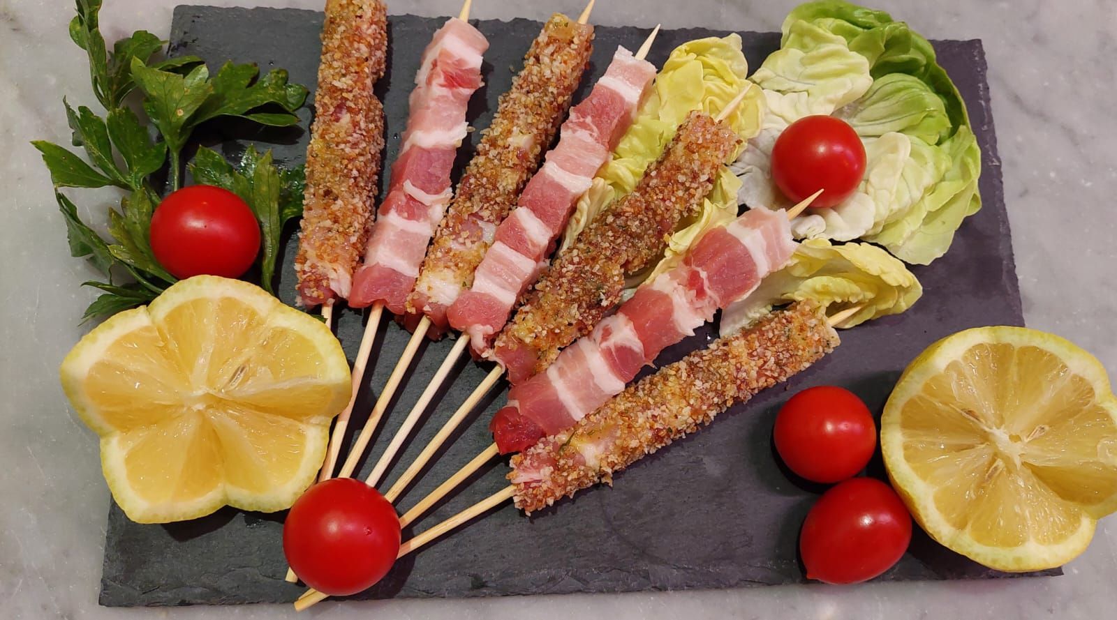 Spiedini di carne avvolti nella pancetta, serviti su un piatto di ardesia con mezzo limone, pomodorini e guarnizione di lattuga.