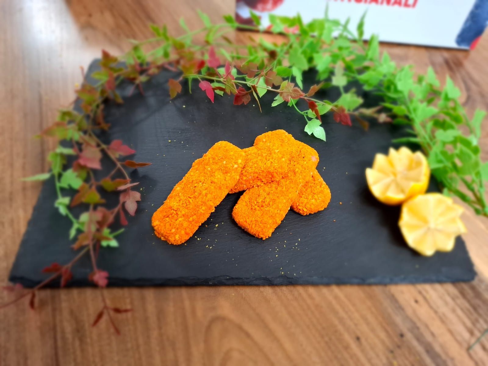 Piatto in ardesia nera con snack fritti arancioni, guarnizione verde e spicchi di limone su un tavolo di legno.