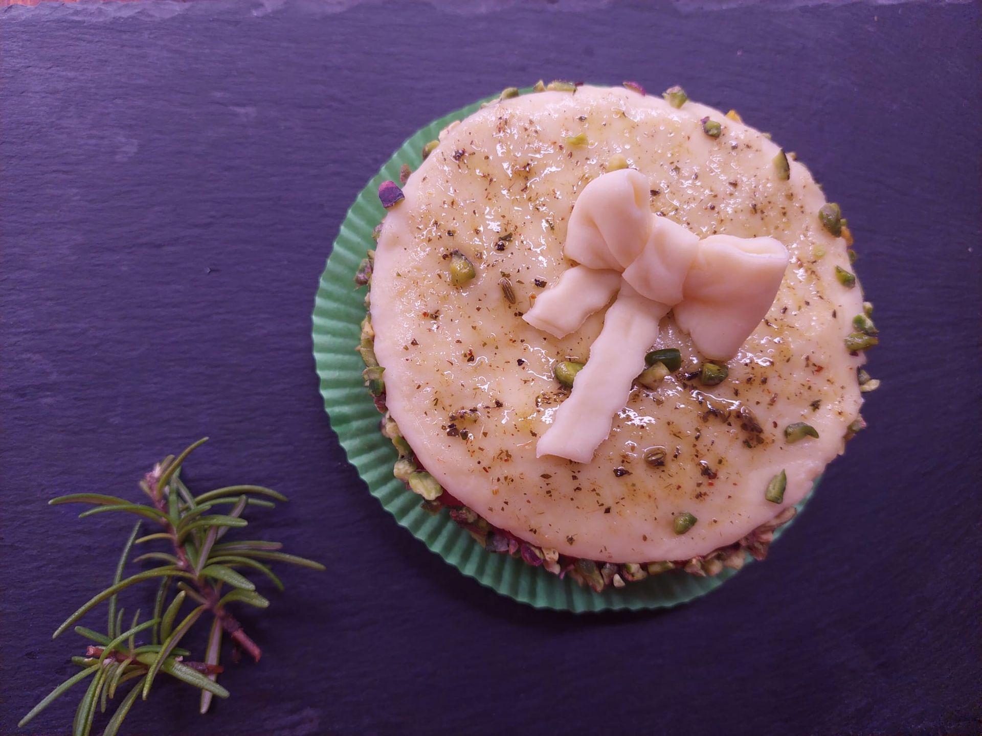 Un cupcake decorato con glassa, sormontato da una rosa rosa su un pirottino verde, accanto a un rametto di rosmarino.