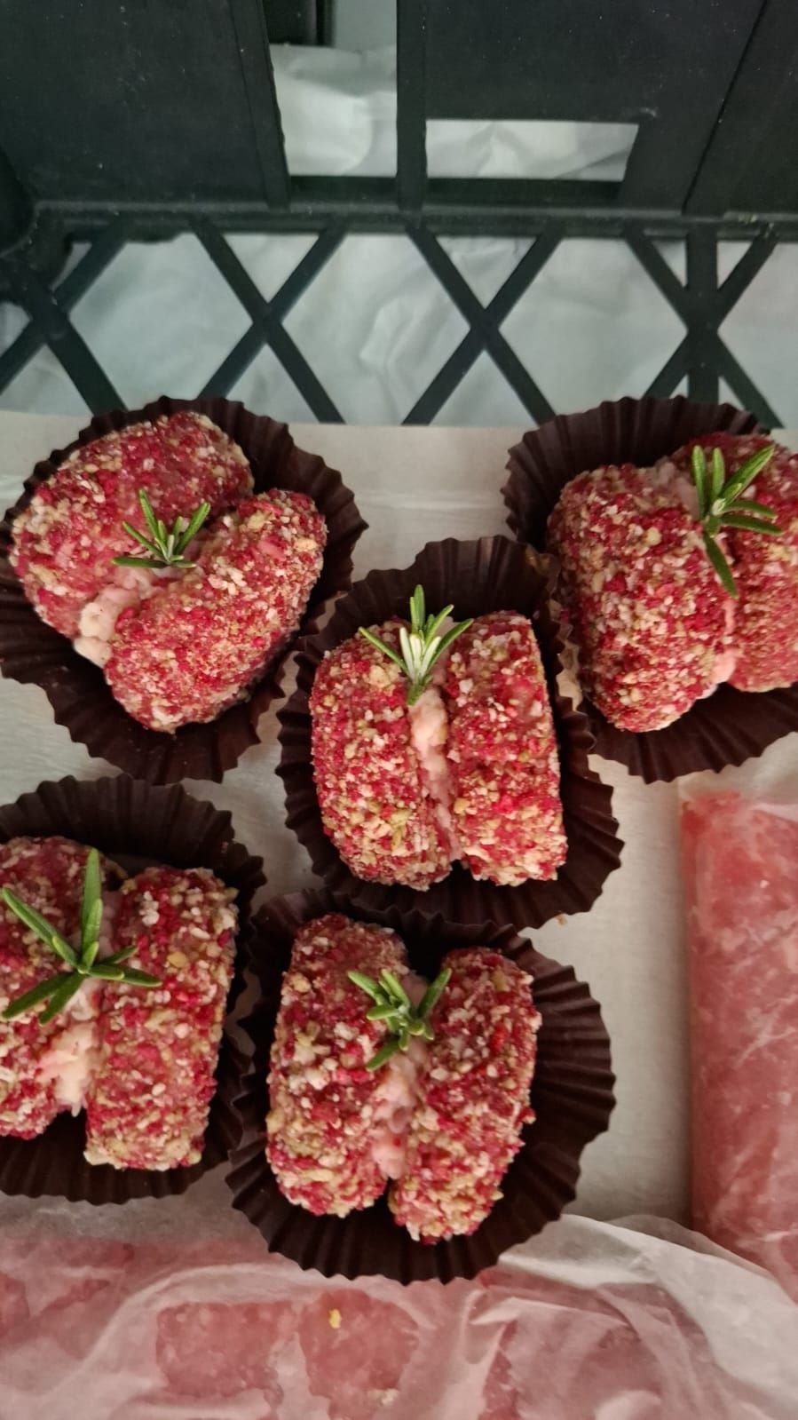Dolcetti rosa ricoperti di zuccherini, disposti su un vassoio in pirottini di carta scura, con una fetta di anguria sulla destra.