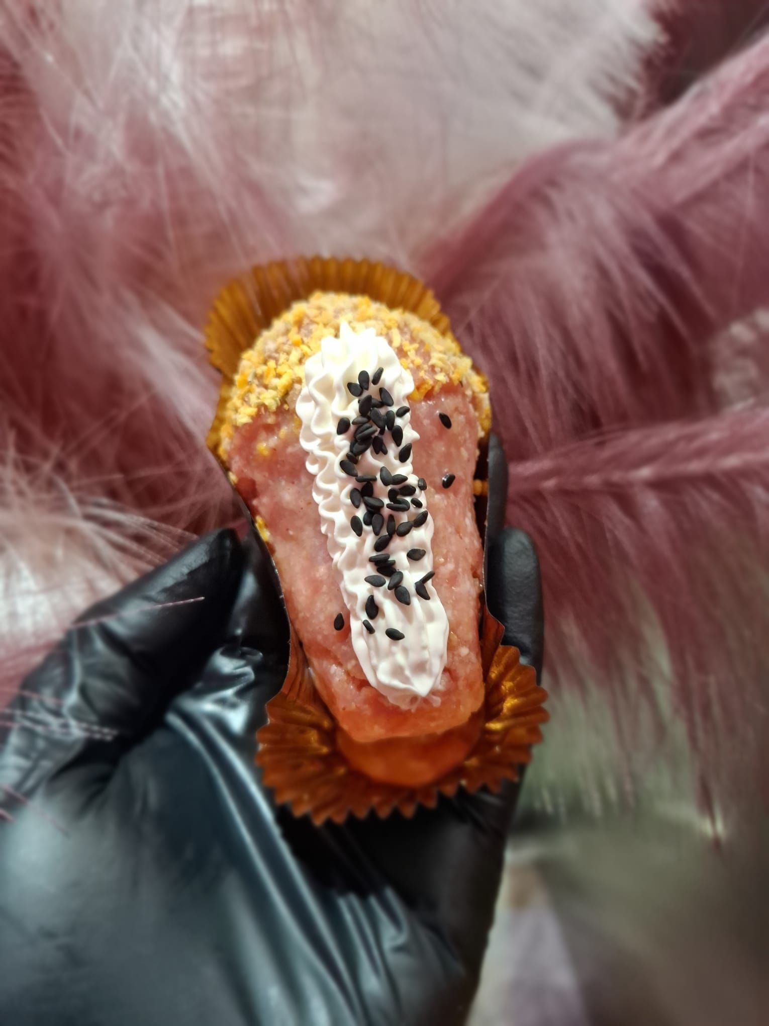 Mano guantata di nero che regge un pasticcino rosa e oro decorato con gioielli, su uno sfondo di pelliccia sintetica rosa.