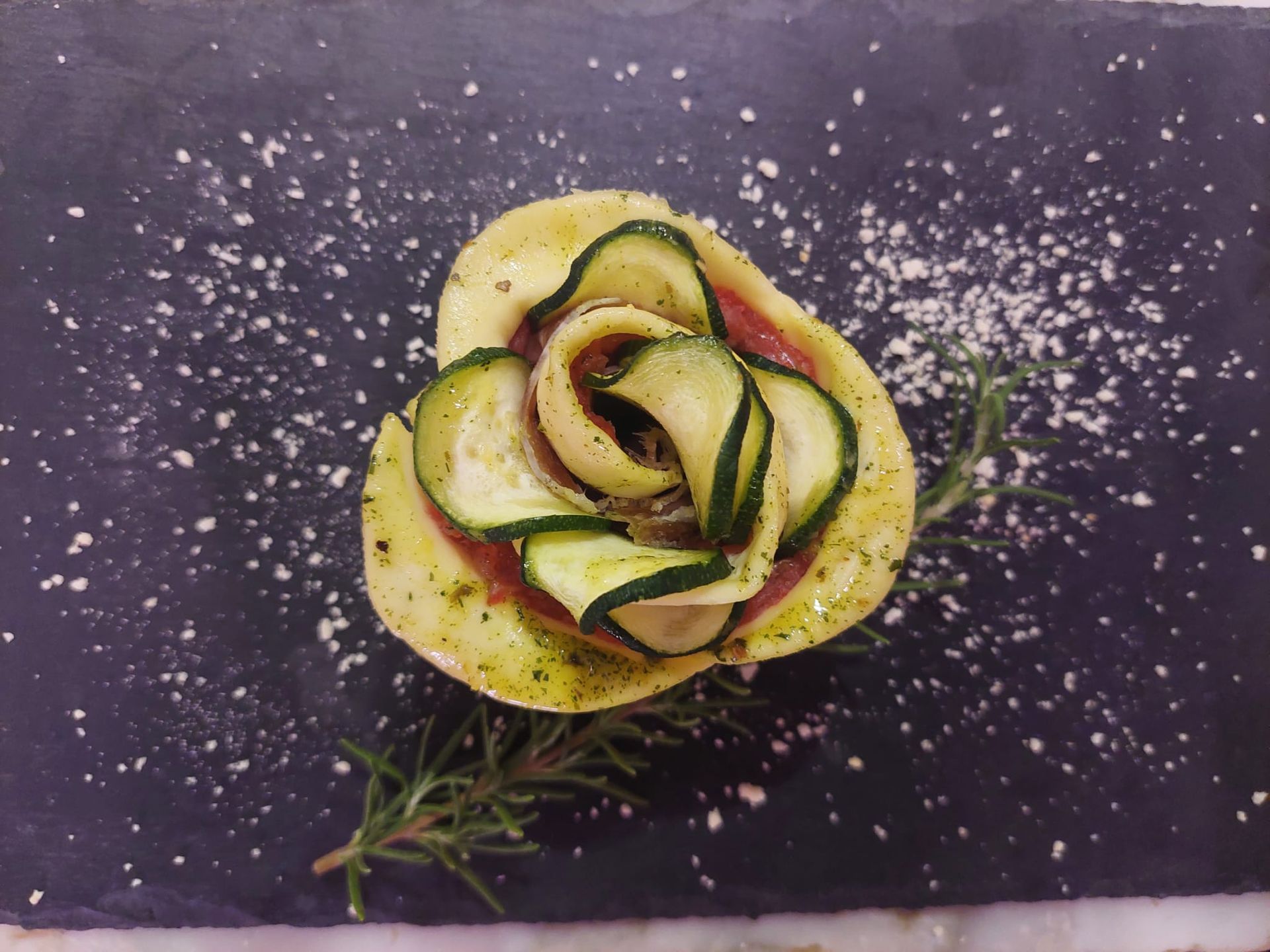 Insalata di verdure a forma di rosa su un piatto scuro, guarnita con erbe aromatiche e cosparsa di spezie.