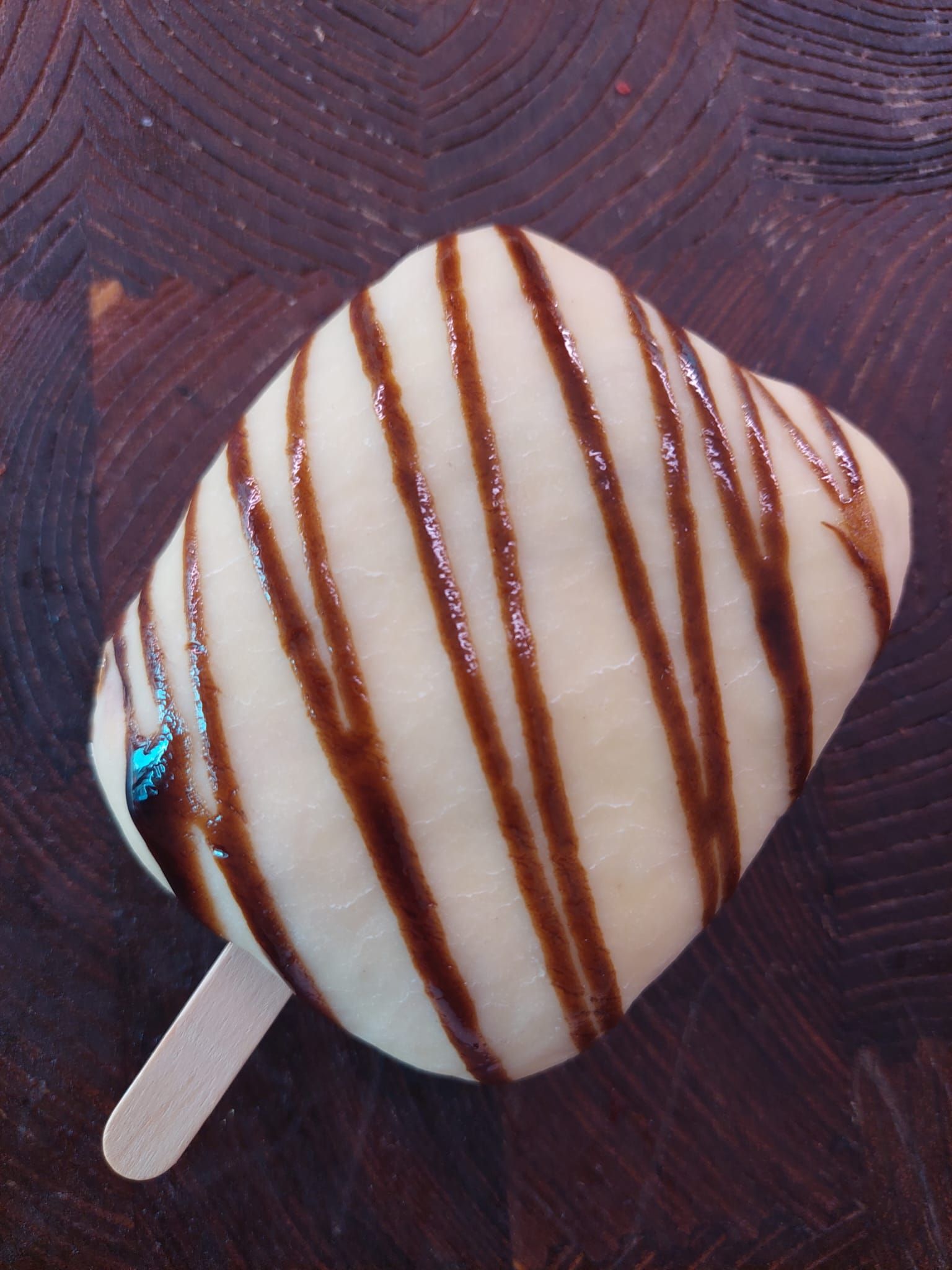 Ghiacciolo bianco con glassa al cioccolato su una superficie di legno, parzialmente avvolto da una piccola etichetta blu.