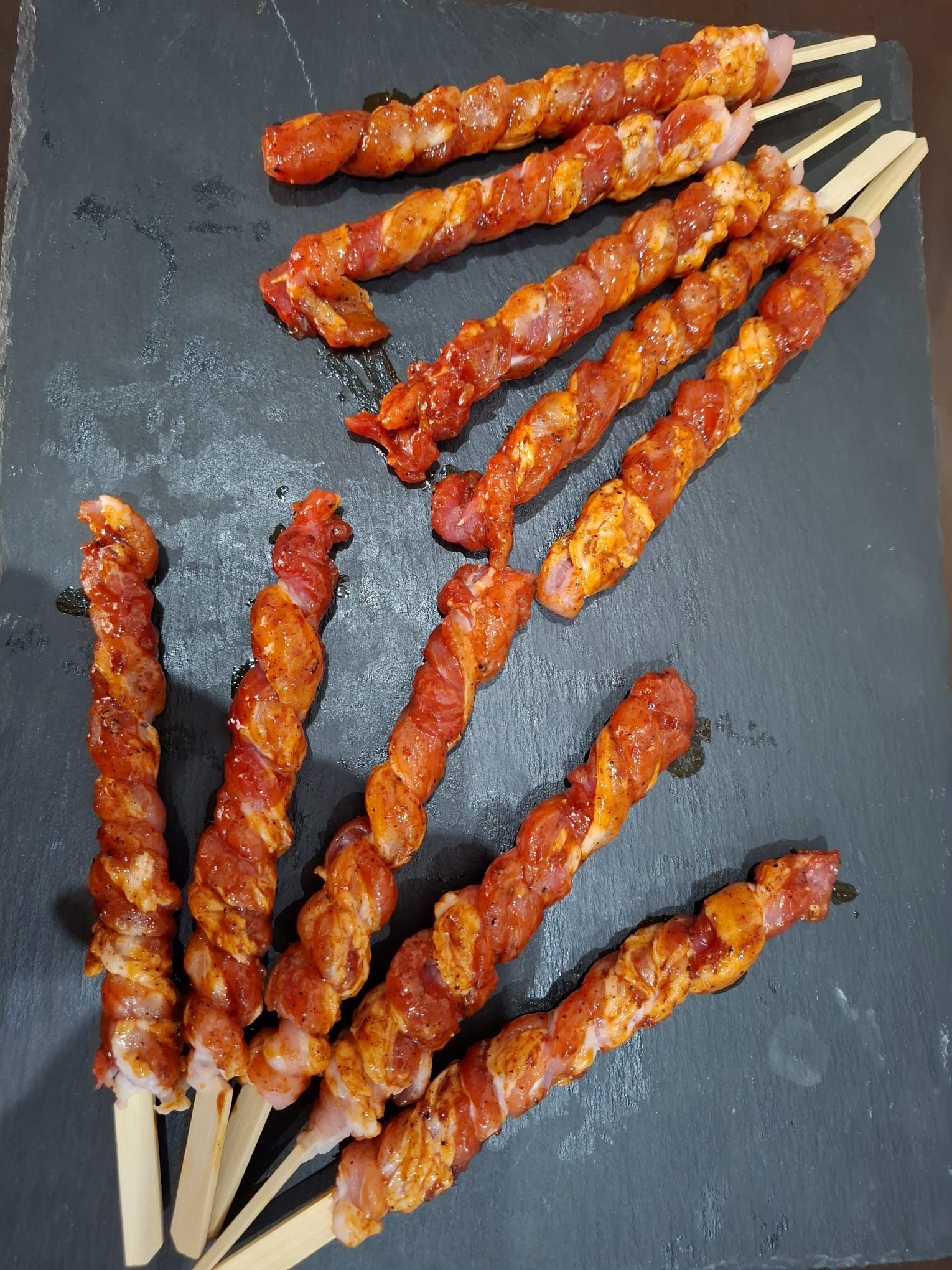 Striscioline di carne grigliata infilzate su uno spiedo di ardesia scura, con una glassa lucida di colore bruno-rossastro.