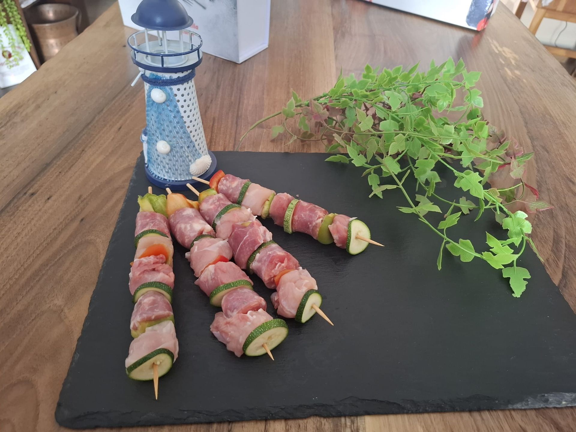 Spiedini di verdure avvolti nella pancetta su un tagliere di ardesia nera con un contenitore a forma di faro blu e verdure nelle vicinanze.