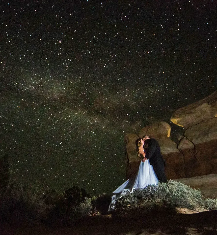 A couple in wedding attire embraces on a rocky hill under a vast, star-filled night sky with the Milky Way visible.