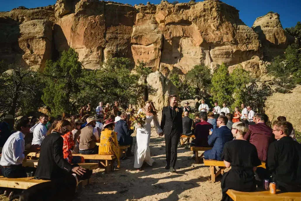 A couple walks hand-in-hand down an aisle of wooden benches at a canyon outdoor wedding, surrounded by guests.