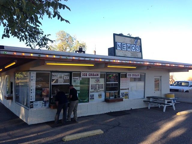 Two people stand at the service window of a white, single-story Nem's ice cream stand on a sunny day.