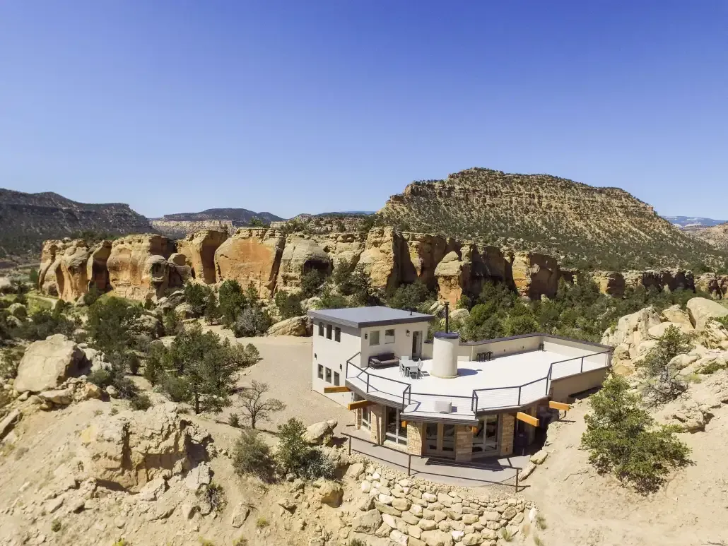 A modern, flat-roofed house built into a desert landscape with rocky cliffs and sparse vegetation under a clear blue sky.